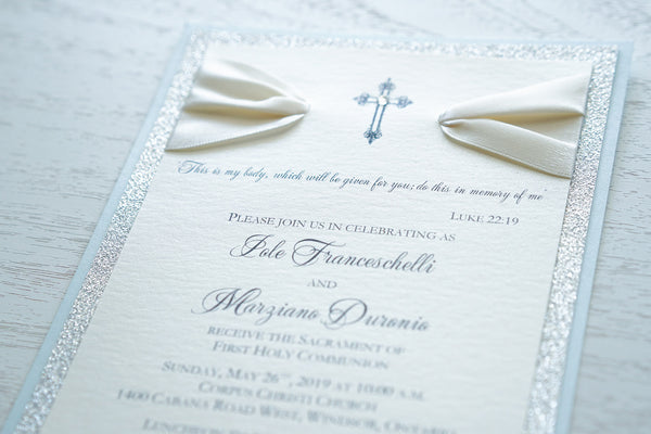 alt=“Elegant glitter First Communion invitation features an ivory pearlescent shimmer card stock on blue pearlescent shimmer and silver glitter card stock, an elegant cross and jewel detail finished with a rich ivory satin ribbon”