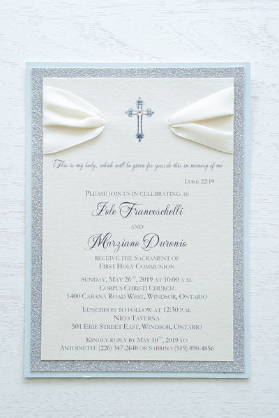 alt=“Elegant glitter First Communion invitation features an ivory pearlescent shimmer card stock on blue pearlescent shimmer and silver glitter card stock, an elegant cross and jewel detail finished with a rich ivory satin ribbon”
