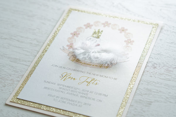 alt=“Whimsical baby shower invitation features white pearlescent shimmer, gold glitter and soft pink card stock with a sweet swan and floral detail with a white feather, a rich pink satin ribbon and jewel accent and a gold glitter crown”