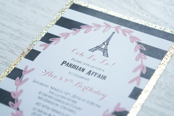 alt="Elegant Parisian birthday party invitation features a matte white stock on a gold glitter card stock, a pink floral leaf frame with a black and white striped background and is finished off with an Eiffel Tower design and jewel detail"
