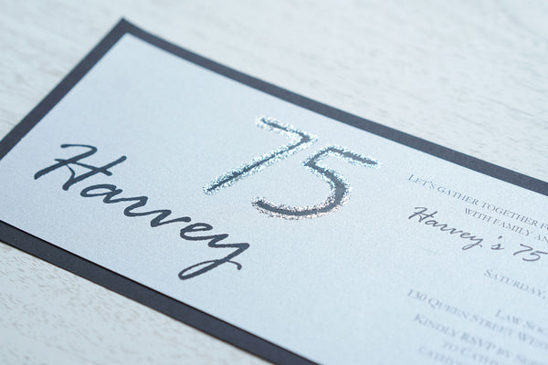 alt=“Modern and fun tea length birthday invitation features a silver pearlescent shimmer card stock on a matte black stock, the honouree’s name in script and their age highlighted with silver glitter sparkle”