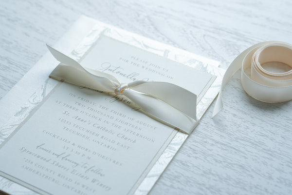 alt="Romantic wedding invitation features ivory matte linen and champagne pearlescent shimmer card stock layered onto a champagne patterned back layer with vintage filigree, scrolls, swirls and etched leaves and is tied together with a a rich ivory satin ribbon"