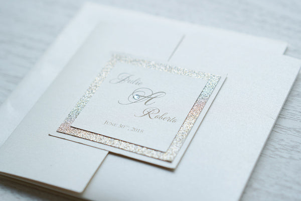alt="Luxurious ivory pearlescent shimmer square pocket fold wedding invitation features an ivory pearlescent shimmer stock on a gold pearlescent shimmer stock, gold font and a coordinating tab with champagne gold glitter stock layer and a monogram/jewel detail to finish it off"