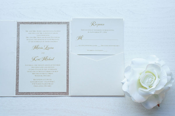 alt="Elegant ivory pearlescent shimmer pocket fold wedding invitation features an ivory pearlescent shimmer stock on a rose gold glitter stock, gold font and a coordinating tab with monogram/jewel detail to finish it off"