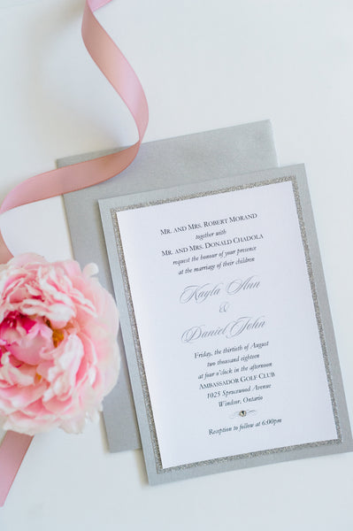alt="Modern wedding invitation features a white pearlescent shimmer card stock layered onto silver glitter and pearlescent shimmer stock layers, a jewel detail and a coordinating belly band with couple's names and wedding date"