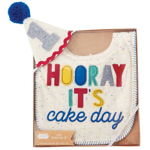 alt=“Confetti speckle cotton knit bib features felt, knit and embroidered Hooray It’s Cake Day sentiment with coordinating confetti speckle covered felt birthday hat with yarn pom-pom topper, ric-rac detail and felt 1 applique”