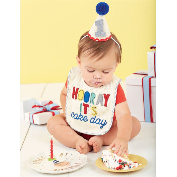 alt=“Confetti speckle cotton knit bib features felt, knit and embroidered Hooray It’s Cake Day sentiment with coordinating confetti speckle covered felt birthday hat with yarn pom-pom topper, ric-rac detail and felt 1 applique”