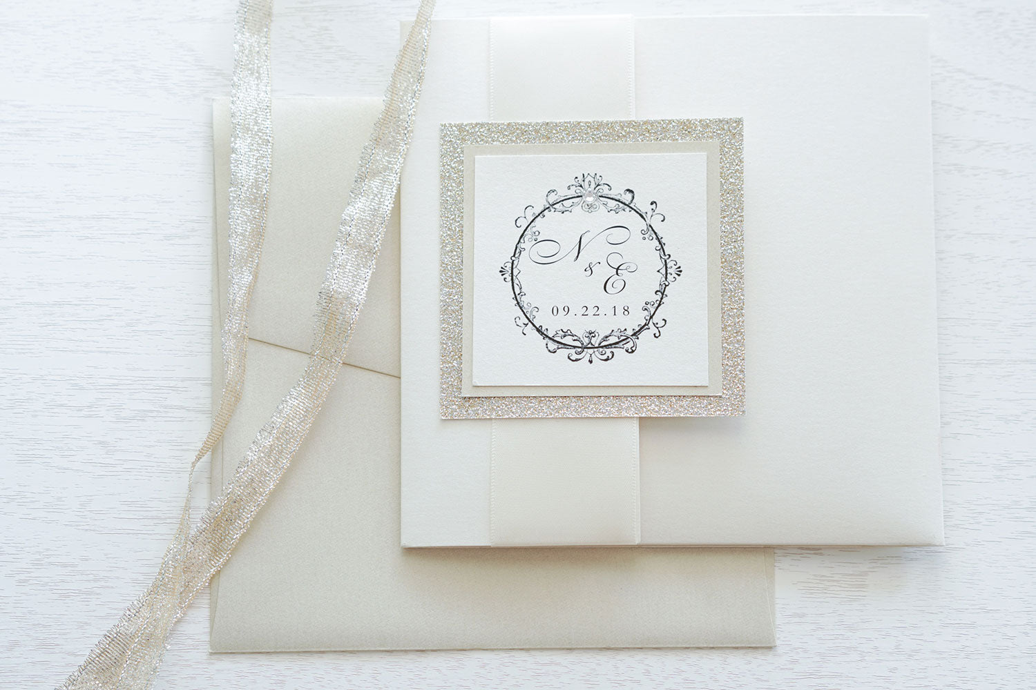alt="Timeless ivory pearlescent shimmer square pocket fold wedding invitation features an ivory pearlescent shimmer stock on a champagne gold pearlescent shimmer stock and is finished on the front with a rich satin ivory ribbon and tab with a champagne gold glitter stock layer and a frame/monogram detail with jewel accent"
