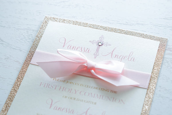 alt=“Sweet glitter First Communion invitation with an ivory pearlescent shimmer card stock on a rose gold glitter stock, a rich pink satin ribbon and bow and an elegant pink cross and jewel detail with the honouree’s name”