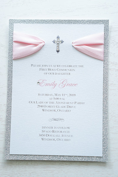 alt="Elegant silver glitter First Communion invitation features a white pearlescent shimmer card stock on a silver glitter stock, a rich pink satin ribbon and an elegant cross and jewel detail"