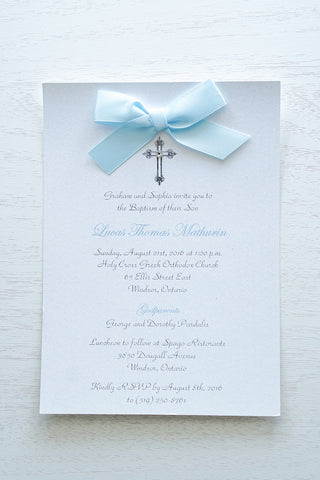 alt=“Classic embellished Baptism invitation features a white pearlescent shimmer card stock, an elegant cross and jewel detail finished with a rich baby blue satin ribbon bow” 