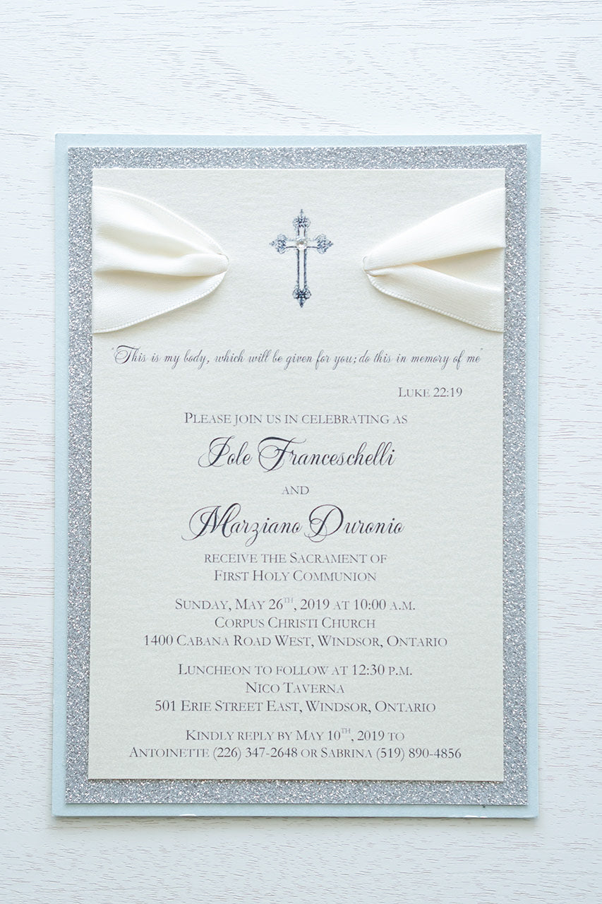 alt=“Elegant glitter First Communion invitation features an ivory pearlescent shimmer card stock on blue pearlescent shimmer and silver glitter card stock, an elegant cross and jewel detail finished with a rich ivory satin ribbon”
