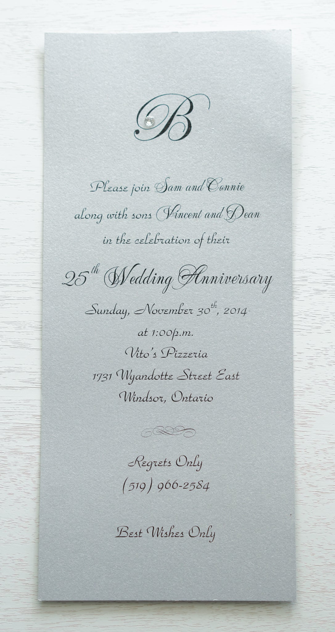 alt=“Simplistic 25th Wedding Anniversary invitation features a silver pearlescent shimmer card stock and an elegant monogram and jewel detail”