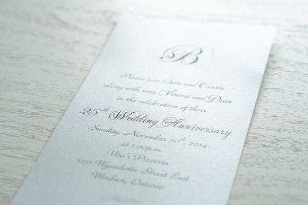 alt=“Simplistic 25th Wedding Anniversary invitation features a silver pearlescent shimmer card stock and an elegant monogram and jewel detail”