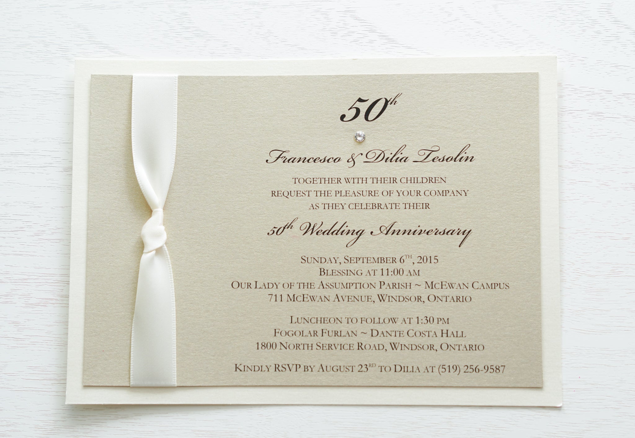 alt=“Classy 50th Wedding Anniversary invitation features a champagne gold pearlescent shimmer card stock on an ivory pearlescent shimmer card stock, an elegant 50th and jewel detail and is finished with a rich ivory knotted ribbon”