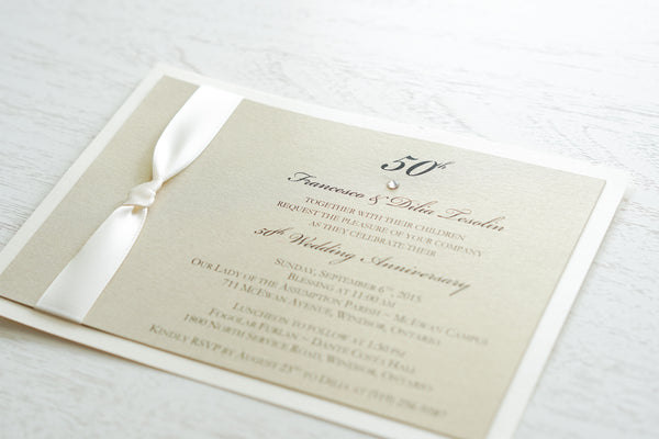 alt=“Classy 50th Wedding Anniversary invitation features a champagne gold pearlescent shimmer card stock on an ivory pearlescent shimmer card stock, an elegant 50th and jewel detail and is finished with a rich ivory knotted ribbon”