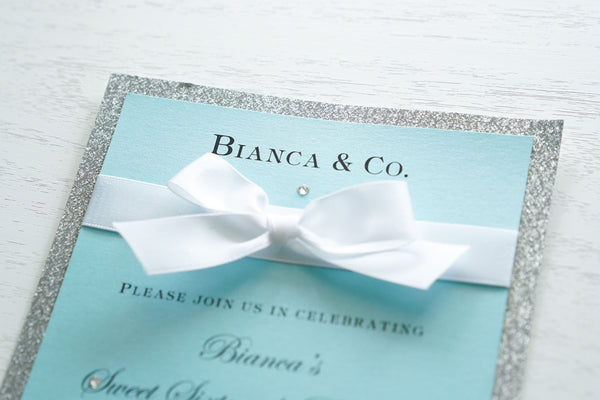 alt="Luxurious Tiffany & Co. inspired birthday invitation features a Tiffany blue pearlescent shimmer card stock on a silver glitter stock, a rich white satin ribbon bow and an elegant jewel detail"