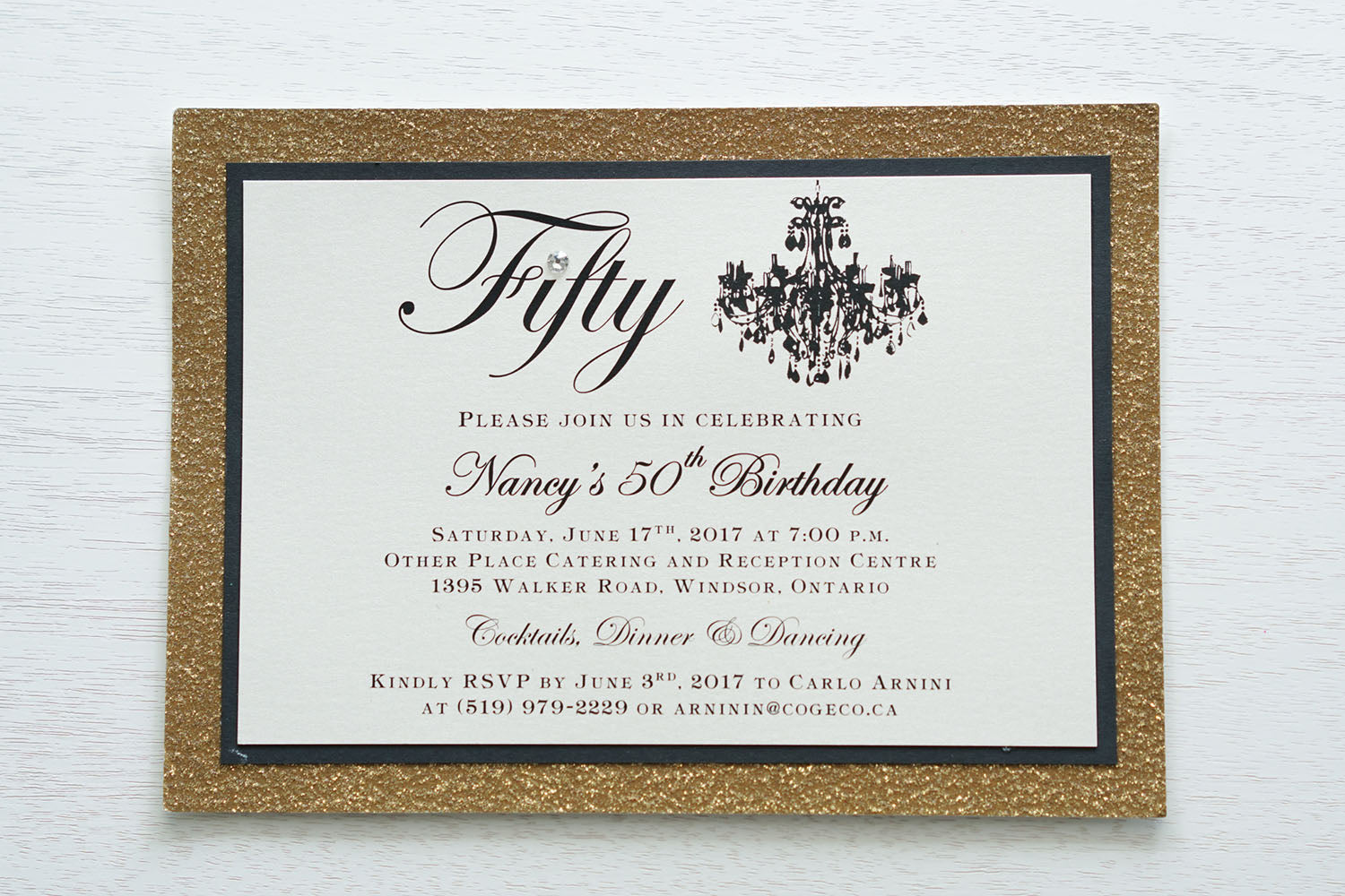 alt="Elegant birthday invitation features an ivory pearlescent shimmer card stock on matte black and gold glitter card stock, a beautiful chandelier image, fifty written in script and is finished with a jewel detail"