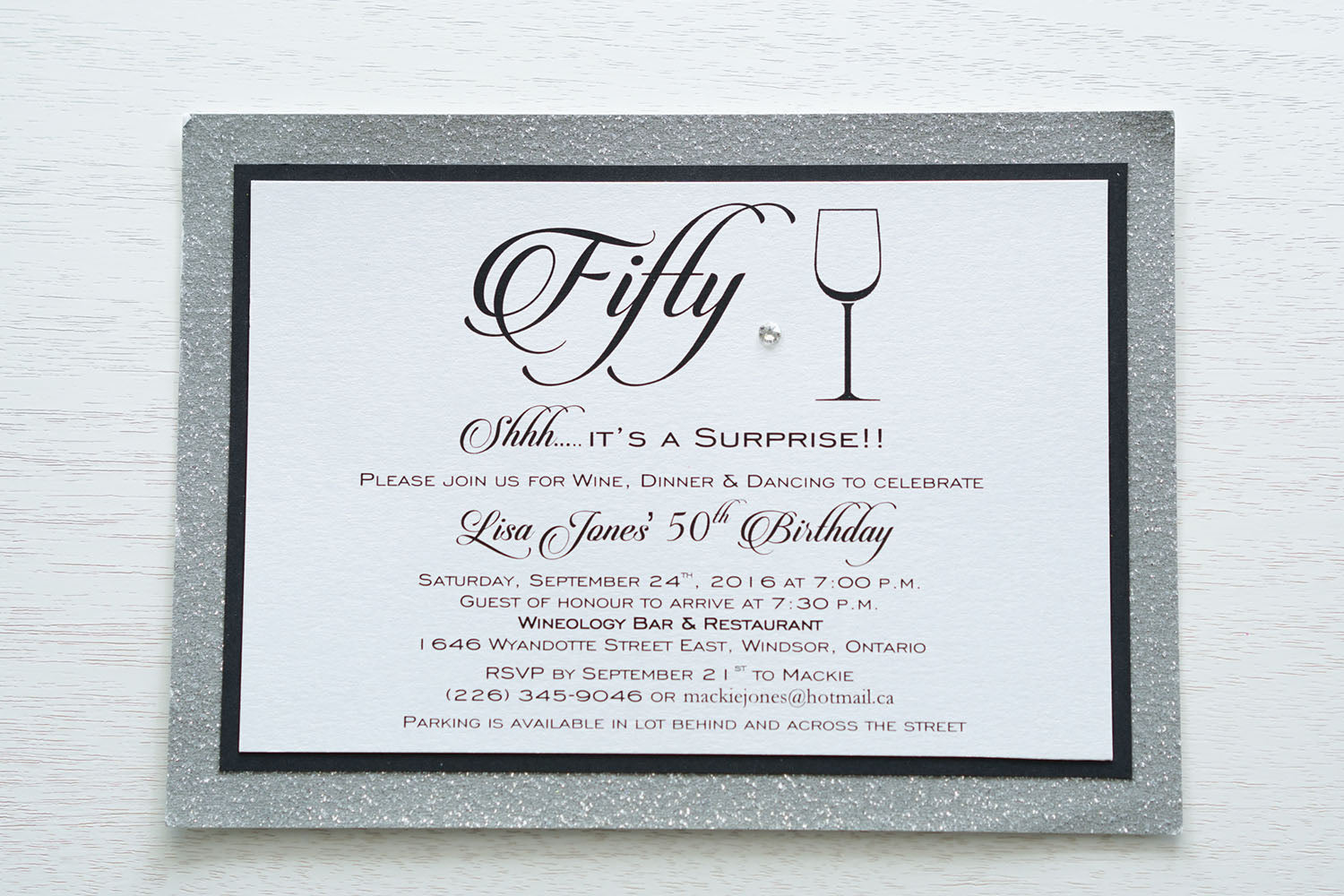 alt="Modern birthday invitation features a white pearlescent shimmer card stock on matte black and silver glitter card stock, a fun wine glass image, fifty written in script and is finished with a jewel detail"