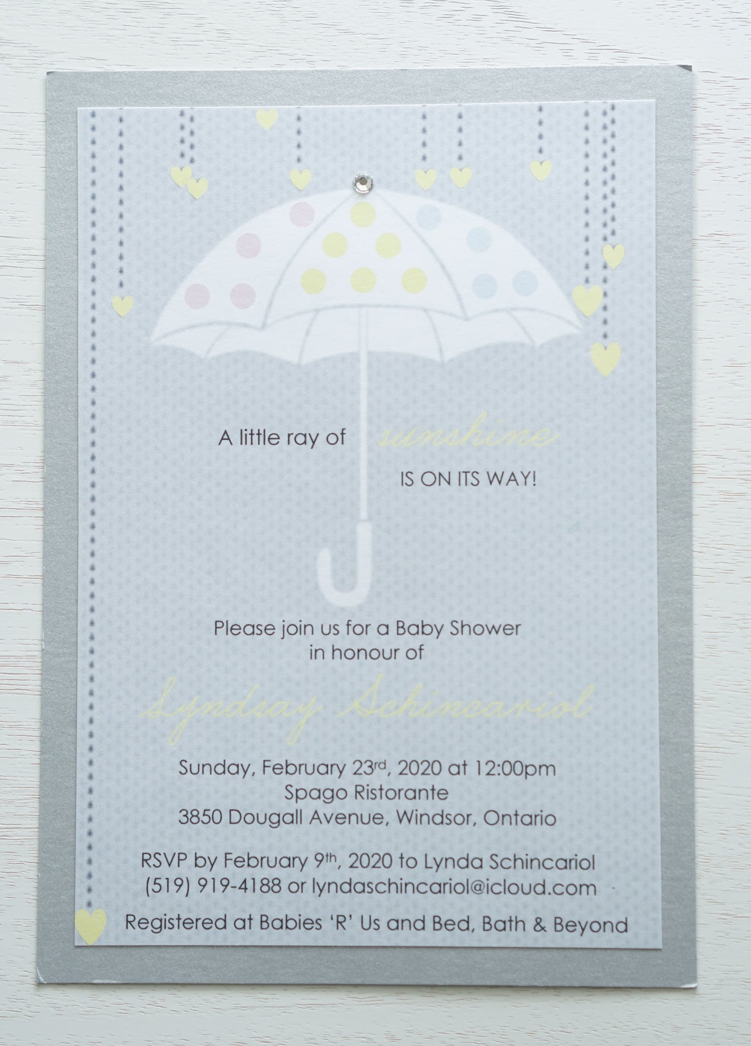alt=“Adorable gender neutral baby shower invitation features a grey background print and an unbrella image with multicoloured polka dots and hearts with a jewel accent and “A little ray of sunshine is on its way”sentiment mounted onto a silver pearlized shimmer card stock layer”