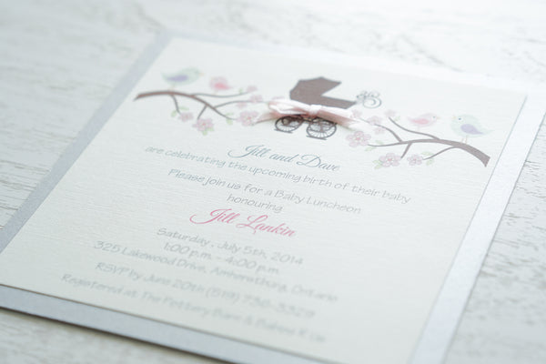 alt=“Whimsical gender neutral baby shower invitation features an ivory linen card stock mounted onto a silver pearlized shimmer card stock, and includes a brown printed pram and tree branches with pastel coloured bird and floral details and is finished with a pink satin ribbon bow detail”