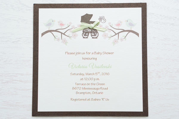 alt=“Whimsical gender neutral baby shower invitation features an ivory linen card stock mounted onto a brown pearlized shimmer card stock, and includes a brown printed pram and tree branches with pastel coloured bird and floral details and is finished with a green satin ribbon bow detail”