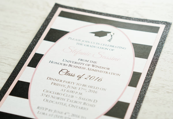 alt="Modern graduation party invitation features a matte white card stock on a pink pearlescent shimmer stock layered on a black glitter stock, a black grad cap and jewel detail and an oval design with a black and white striped pattern to finish it off"