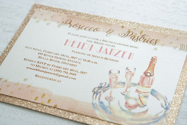 alt="Glam glitter Bridal Shower invitation features an ivory pearlescent shimmer card stock on a rose gold glitter stock, an elegant blush pink watercolour border, a prosecco and macaron design and jewel detail to finish it off"