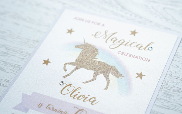 alt="Magical birthday party invitation features a white pearlescent shimmer stock on a light purple pearlescent card stock, an elegant faux glitter unicorn and watercolour rainbow design, a purple banner and gold stars finished with a jewel detail"