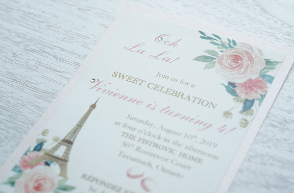 alt="Sweet Parisian birthday party invitation features a matte ivory linen stock on a blush pink pearlescent shimmer card stock, an elegant gold Eiffel Tower, a pink watercolour floral and French macaron design and is finished with a jewel detail"