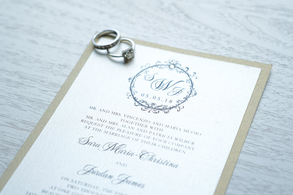 alt="Timeless wedding invitation features an ivory pearlescent shimmer card stock on a gold pearlescent shimmer stock, an elegant script font and a round frame design with a monogram/jewel detail"