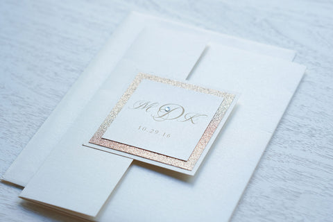 alt="Elegant ivory pearlescent shimmer pocket fold wedding invitation features an ivory pearlescent shimmer stock on a rose gold glitter stock, gold font and a coordinating tab with monogram/jewel detail to finish it off"