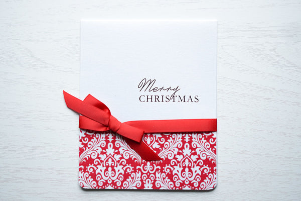 alt=“This elegant card features a white pearlescent shimmer card stock, “Happy Holidays” or “Merry Christmas” printed sentiment in pretty script writing with a red damask pattern and is finished with a rich satin red ribbon bow tied around the card” 