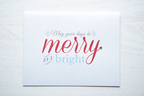 alt="This modern card features a white pearlescent shimmer card stock, “May your days be merry and bright” printed sentiment in grey, red and blue writing and is finished with a jewel detail"
