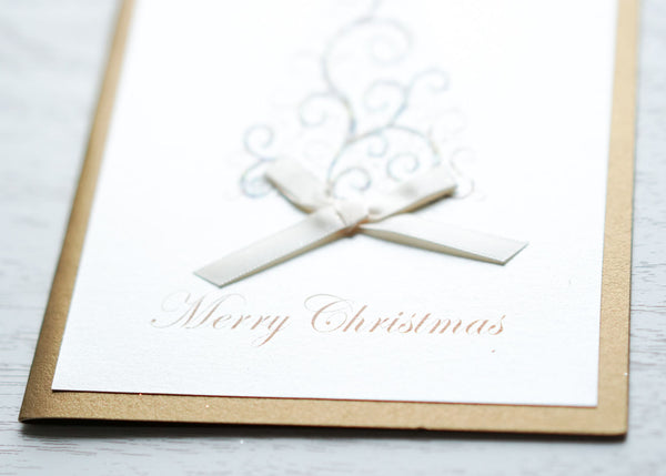 alt="This elegant card features an ivory and antique gold pearlescent shimmer card stock, a gold printed swirl Chirstmas tree with glitter dust, jewel and a rich satin ivory ribbon bow and “Happy Holidays” or “Merry Christmas” printed below in pretty gold script writing"