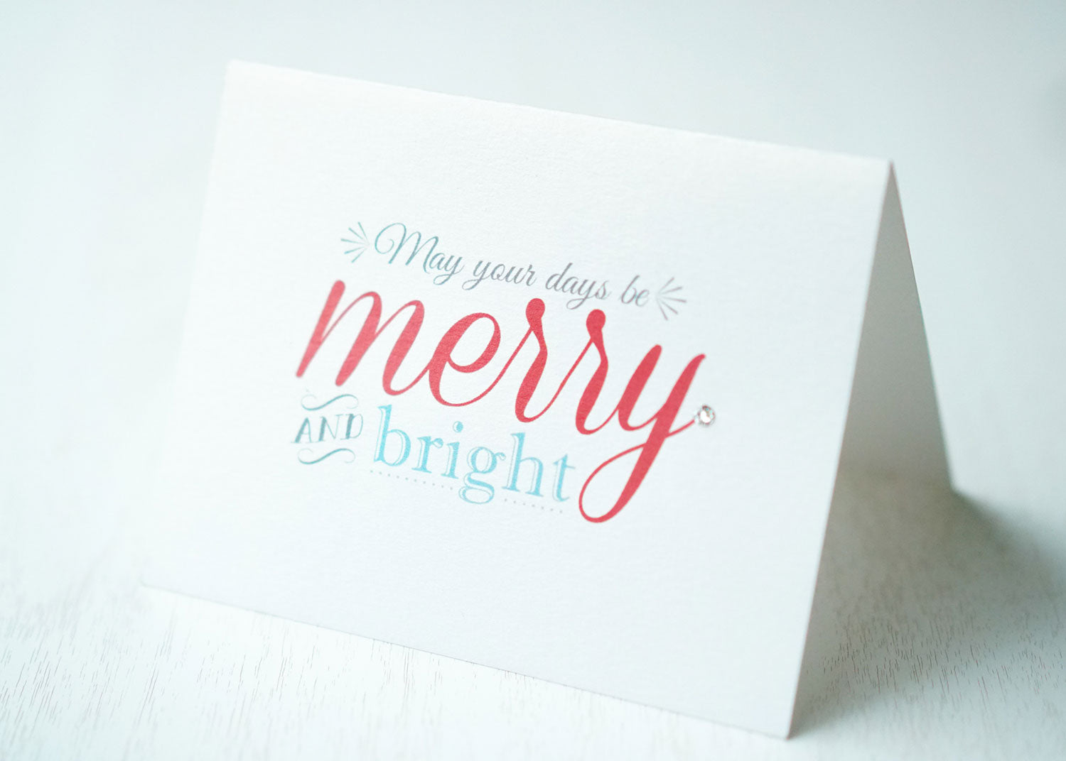 alt="This modern card features a white pearlescent shimmer card stock, “May your days be merry and bright” printed sentiment in grey, red and blue writing and is finished with a jewel detail"