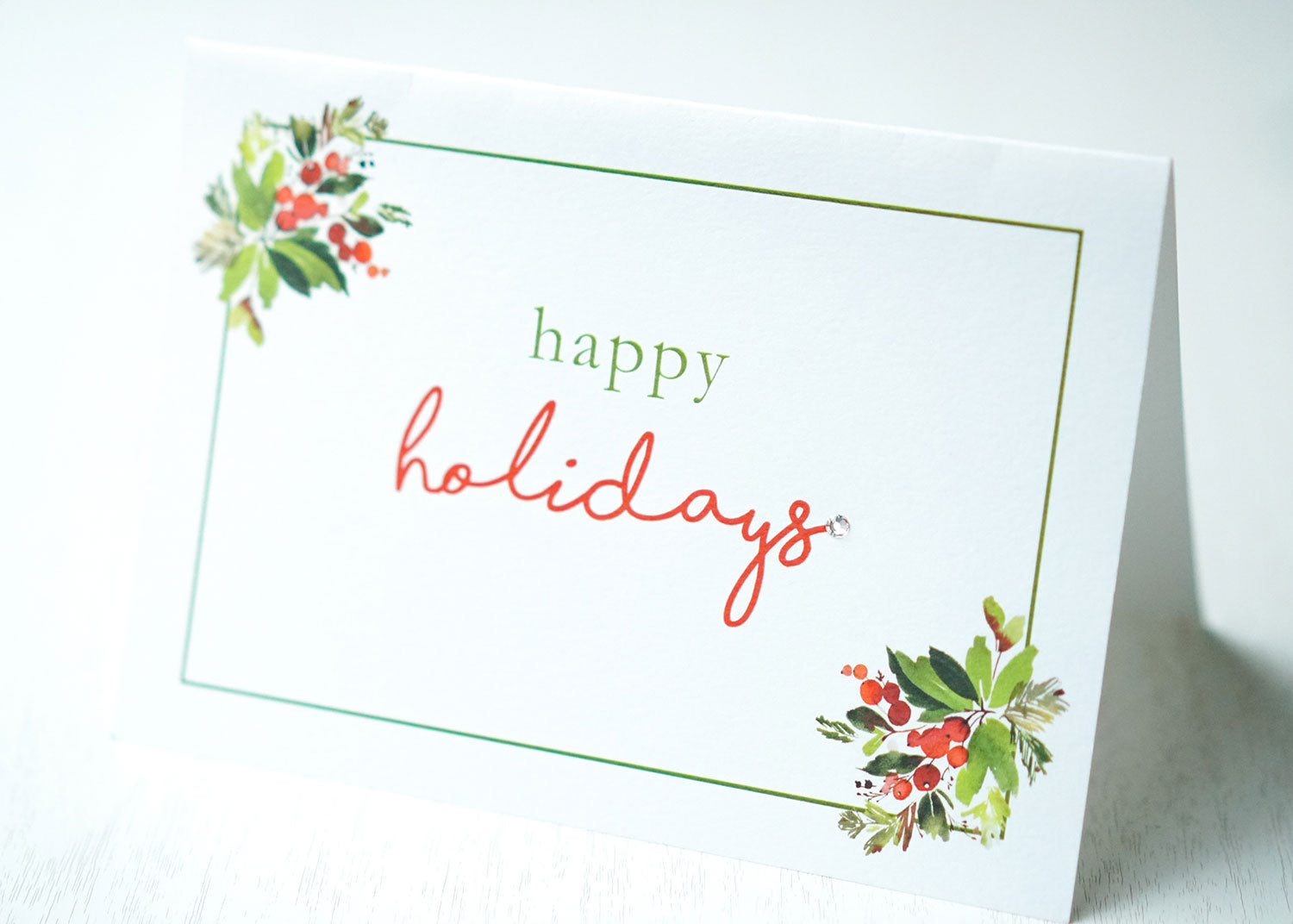 alt="This gorgeous Christmas card features a white pearlescent shimmer card stock, “Happy Holidays” or “Merry Christmas” printed sentiment in pretty script writing with a jewel detail and is framed by a green border, leaves and berries"