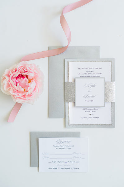 alt="Modern wedding invitation features a white pearlescent shimmer card stock layered onto silver glitter and pearlescent shimmer stock layers, a jewel detail and a coordinating belly band with couple's names and wedding date"